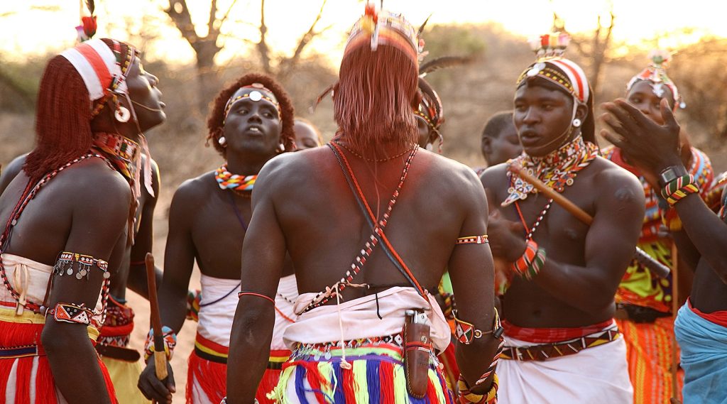 Samburu men dressed in traditional attire singing and performing, with vibrant colors and cultural decorations.