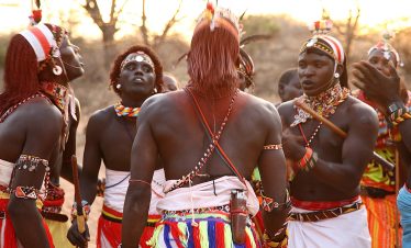 Samburu men dressed in traditional attire singing and performing, with vibrant colors and cultural decorations.