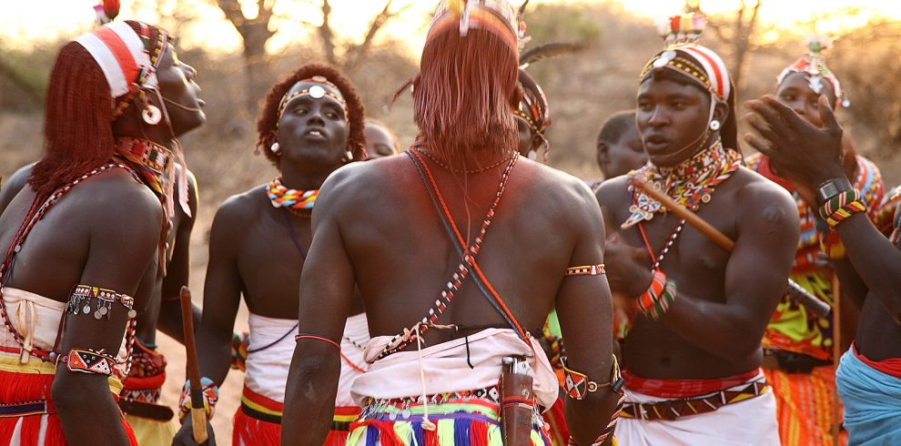 Samburu men dressed in traditional attire singing and performing, with vibrant colors and cultural decorations.