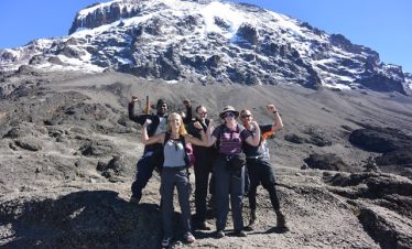 A group of tourists standing together, celebrating and taking a photo on Mount Kilimanjaro with a backdrop of clouds and mountainous scenery.