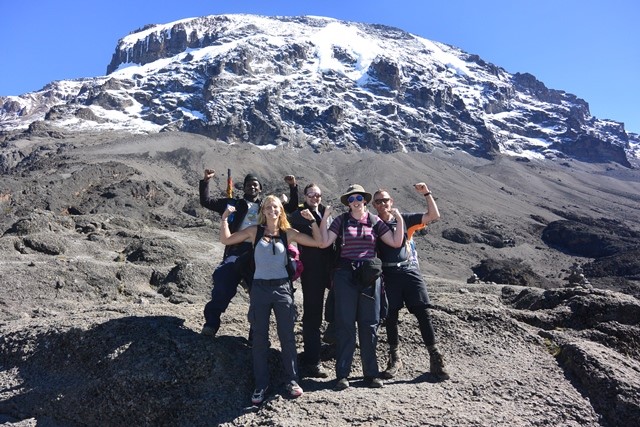 A group of tourists standing together, celebrating and taking a photo on Mount Kilimanjaro with a backdrop of clouds and mountainous scenery.