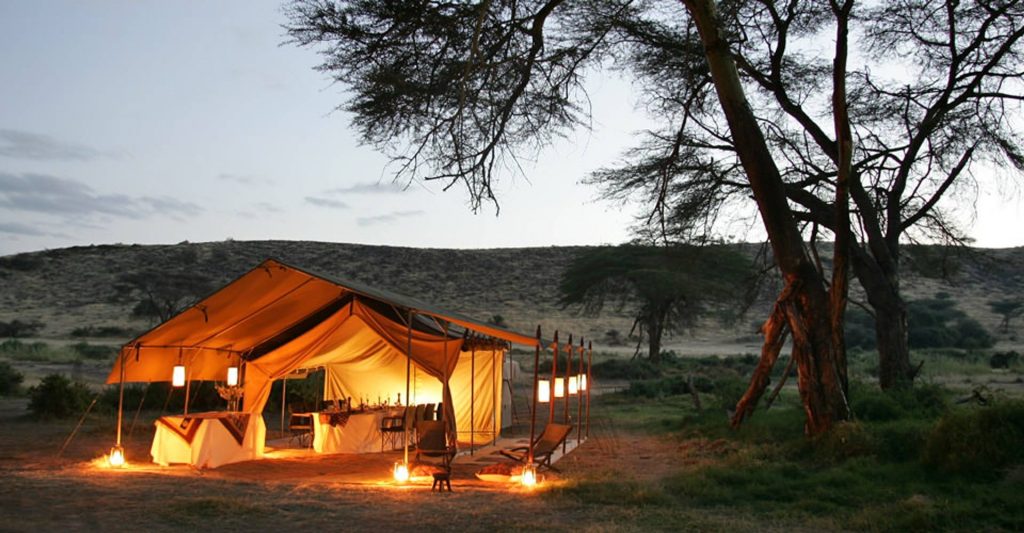 A camp setup in the Kenyan wilderness with tents and campfire surrounded by nature.