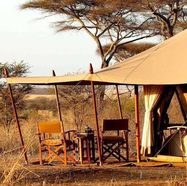 A tent camp set up in the Tanzanian landscape, featuring safari tents and a campfire with a backdrop of savanna.